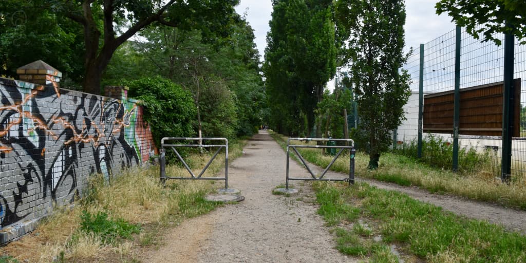 A grassy trail in Berlin with a lovely summer vibe, there is some tasteful graffiti to help color the fence on the left side. Photo by Xe Iaso, 2023.